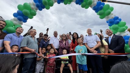Supervisor Joe Baca, Jr., Senator Eloise Gómez Reyes, San Bernardino City Mayor Helen Tran, Congressman Pete Aguilar, Rialto Mayor Joe Baca, Councilmember Treasure Ortiz, Redlands Mayor, SBCTA President, and community members gather with children under a green, blue, and white balloon arch for a ribbon-cutting ceremony.