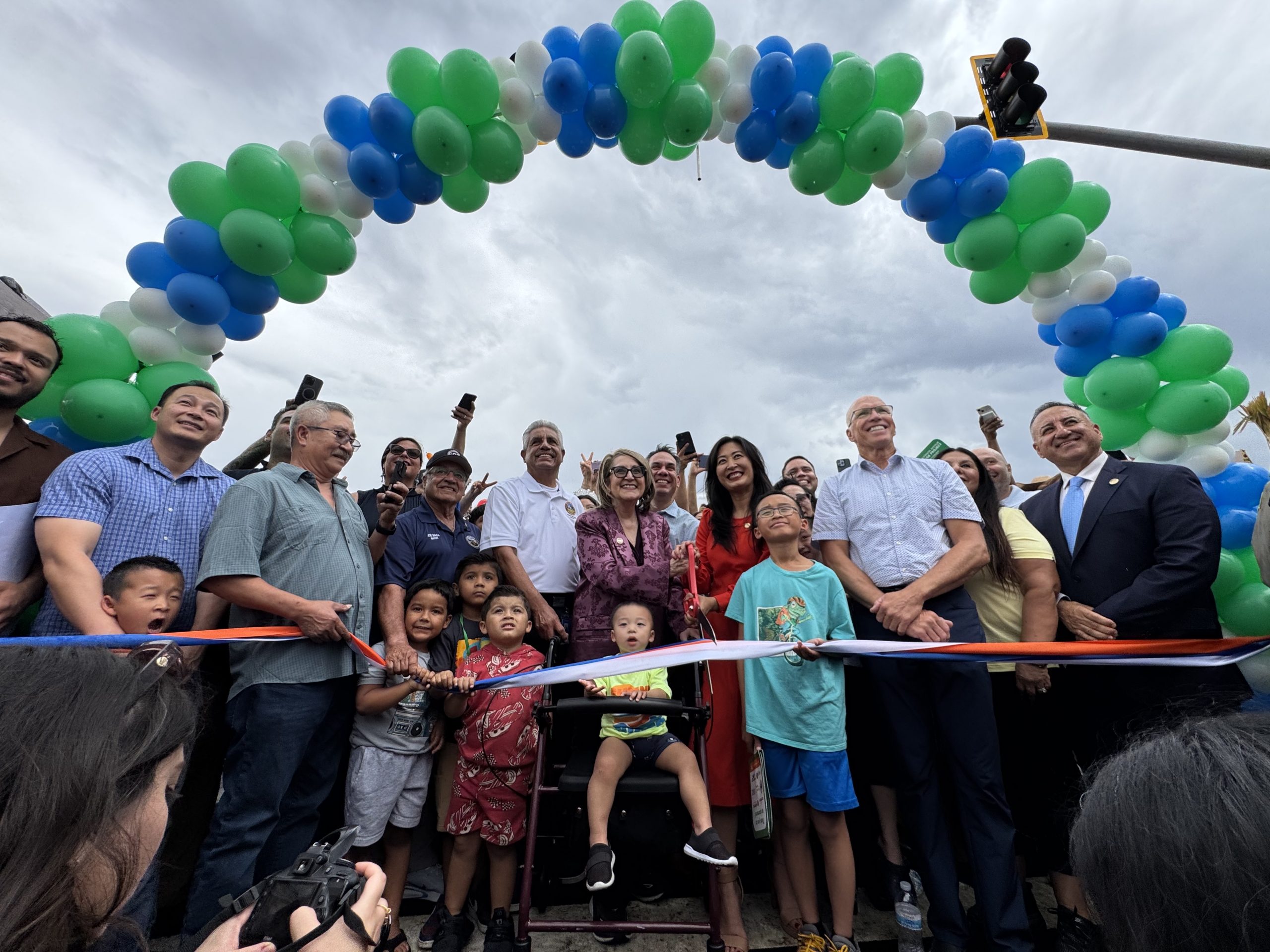 Supervisor Joe Baca, Jr., Senator Eloise Gómez Reyes, San Bernardino City Mayor Helen Tran, Congressman Pete Aguilar, Rialto Mayor Joe Baca, Councilmember Treasure Ortiz, Redlands Mayor, SBCTA President, and community members gather with children under a green, blue, and white balloon arch for a ribbon-cutting ceremony.